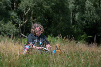 A woman with curly dark blonde hair sits in long grass drawing