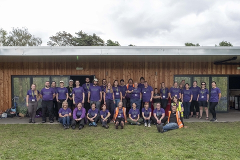 A group of people in purple tshirts stood and sat outside a wooden clad building