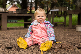 A blonde female toddler sits on the ground covered in dirt in a pink jump suit and yellow boot covers