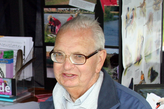An elderly man sits next to a table holding a mug. He wears glasses, a white rugby style top and a blue waterproof coat