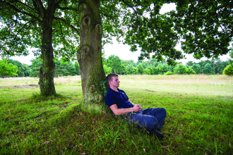 A man with blonde hair sits leaning against a tree in wood pasture, he has his legs crossed and hands clasped lightly together and looks peaceful.