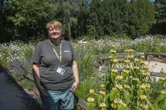 A woman with short blonde hair smiles at the camera, she wears a grey tshirt and stand in a sunny garden with wildflowers growing