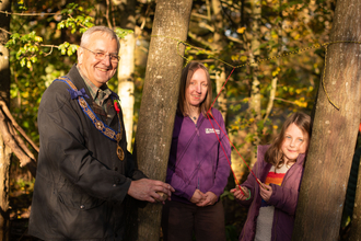 A man with grey hair and classes smiles at the camera, next to him is a blonde long haired woman in a purple jumper and a young girl with blonde hair and a rainbow top. They stand between trees, holding wooden discs hanging on strings