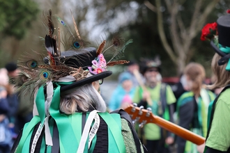 A woman with dark grey hair wears a black hat covered in colourful feathers and a cape with lots of green ribbons