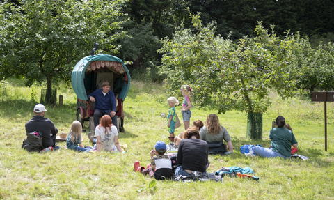 A man sits next to an old fashioned gypsy wagon in front of groups of people and children in a green orchard setting with apple trees dotted among the grass. The people sit on the floor watching the man and a sign says storytelling pointing towards the man.