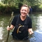A woman with blonde hair tied up smiles the camera as she stands waist deep in a river, she holds a large stick in the water and wears black waders and a black top.