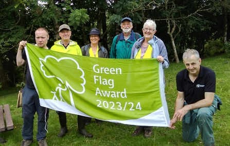 A group of 6 people all hold up a large green flag and smile. The flag has text that reads Green Flag Award 2023/24. They stand on a grassy area with woodland behind them.