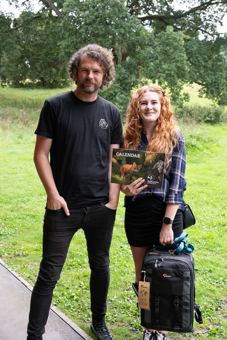 A tall man with dark curly hair wearing black jeans and tshirt stands with a woman with long curly ginger hair, she wears a check shirt, black skirt and holds a calendar with a photo of a fox on the cover, and a big camera bag