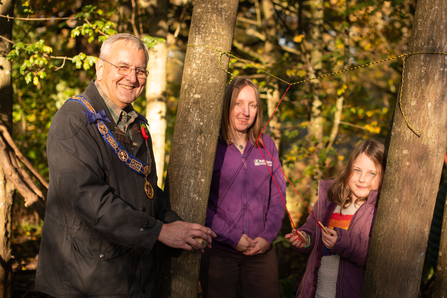 A man with grey hair and classes smiles at the camera, next to him is a blonde long haired woman in a purple jumper and a young girl with blonde hair and a rainbow top. They stand between trees, holding wooden discs hanging on strings