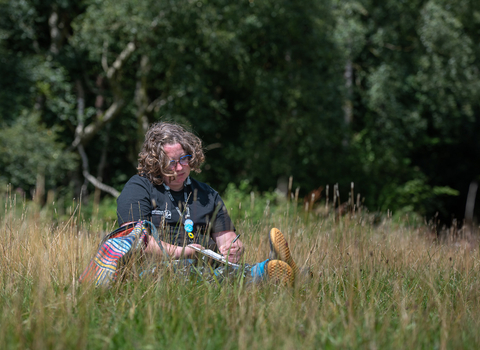 A woman with curly dark blonde hair sits in long grass drawing