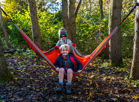 A boy with blonde hair sits in a hammock smiling with another boy in a woolley hat behind him