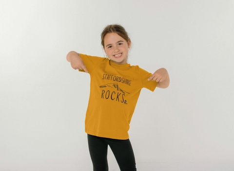 A young girl with light brown hair smiles and points with both hands to her t-shirt which is ochre yellow and says Staffordshire Rocks