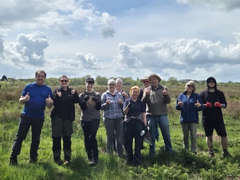 A large group of people smiling at the camera giving thumbs up! They stand in an outdoor area with a wetland behind them and cloudy sky overhead