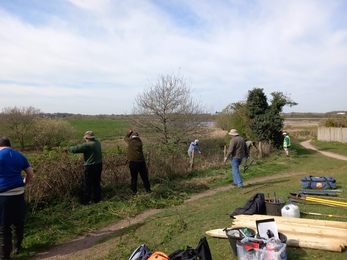 A group of people working to replace/repair fencing at a nature reserve. They sky above is blue with light cloud. A pile of wood and tools lies on the ground to the right and the fence borders a path.