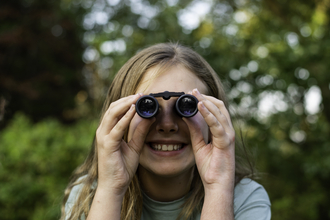 A person peers at the camera through binoculars. She has long straight hair and a big smile. Photo by Evie and Tom Photography.