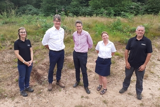 A group of five people stood on sandy area with greenery and trees behind them