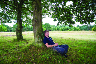 A man with blonde hair sits leaning against a tree in wood pasture, he has his legs crossed and hands clasped lightly together and looks peaceful.
