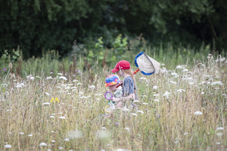 Two children in brightly coloured caps carry a large net as they hunt bugs in a meadow full of long grass