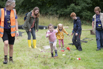 Three children run through an obstacle course with three adults looking on, they are in field with green grass and trees in the distance