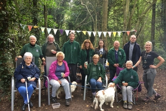 A group of woman and men stand and sit in two rows in a woodland setting with bunting strung up between the trees