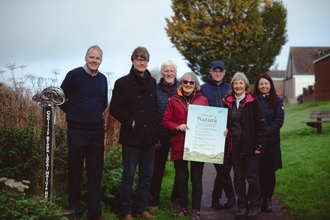 A group of people smile at the camera while holding a Plan for Nature sign, there is grass and trees behind them