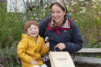 A woman and child sit in a wildflower garden smiling, the woman holds a wildchild paper bag and wears a head scarf, the child holds an icecream