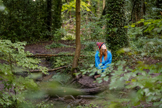 A girl with long ginger hair sits in a green forest surrounded by trees