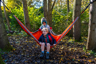 A boy with blonde hair sits in a hammock smiling with another boy in a woolley hat behind him