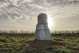 A large stone statue of a women with three faces stands on grass with trees in the distance, the sun is behind the statue with wispy clouds