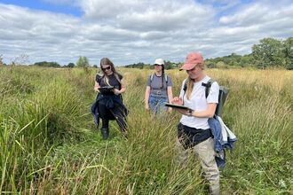 Three woman stand in a field of long grass with clipboards, two wear caps and the sky is blue with wispy clouds and trees in the distance