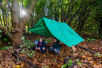 A bright green tarpaulin hangs over a line creating a den in a wooded area. Two boys crouch underneath the den roof and smile. A sunburt shines through the trees and lots of brown leaves cover the floor.