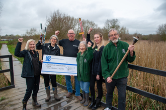 A group of people smile and raise their hands and various hand tools in the air. They hold a large cheque with Co-op on it. They stand on a board walk with reeds in the background and the sky is cloudy.