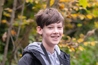 A teenage boy with short brown hair sits on a bench with a camera round his neck. He smiles at the camera. He wears a grey hoodie and pale blue jeans with a black waterproof coat.