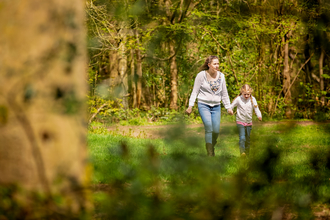 A woman a young girl walk hand in hand through a woodland along a path surrounded by grass. The woman has long hair worn half tied back and wears a grey top and blue jeans, the girl has blonde hair and also wears a grey top and blue jeans.