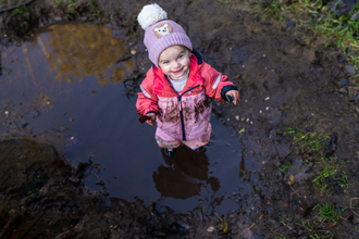 A young girl toddler stands in a muddy puddle - she wears a pink waterproof onsie suit and woolen bobble hat