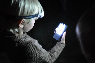 A young boy looks at a bat detector under the light of a headlamp.