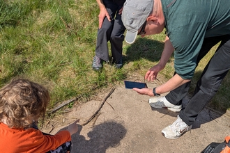 A woman stands watching as a man uses his phone to take a photo of something on the floor that a small child points at