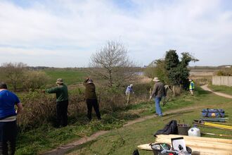 A group of people working to replace/repair fencing at a nature reserve. They sky above is blue with light cloud. A pile of wood and tools lies on the ground to the right and the fence borders a path.