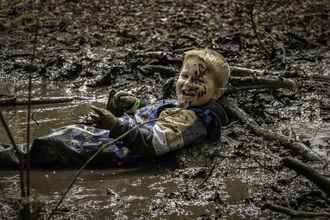 A boy lies in mud/water and has it splattered over his face as he smiles