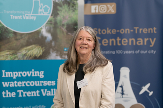 A woman with shoulder length dark grey hair smiles at the camera. She wears a beige jacket and black top and stands in front of two blue banners with text reading "improving watercourses the Trent Valley and Stoke-on-Trent Centenary