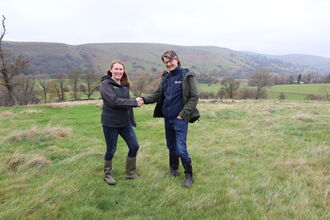 Photograph of Charlotte Harris, CEO Cheshire Wildlife Trust smiling and shaking hands with Julian Woolford, CEO Staffordshire Wildlife Trust both standing facing the camera on green grass with green hills and a grey sky