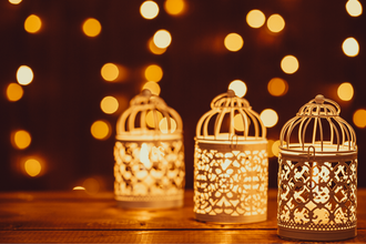 Three Christmas lanterns on a table with fairy lights hanging in the background.