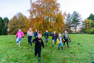 A group of children, both girls and boys, run down a hill on grass in front of an autumnal tree. They all look happy and excited
