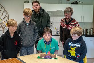 A woman in a green tshirt with short cropped dark brown hair blows out candles on a cake. Two men stand behind her and 3 boys of various ages, all with the same bowl cut blond hair join in to blow out the candles. There's a kitchen area behind them.