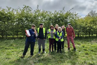 A group of people (men and women) stand together in a field infront of a hedgerow. They wear high vis vests