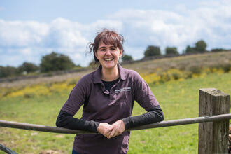 A woman stands resting her arms on a farm gate with a field behind her. She is aged in her 40s with dark brown hair worn up and wears a black top. She smiles and looks content. The sky is blue with fluffy clouds.