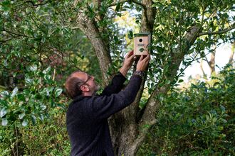 A man in a long sleeved top careful positions a bird box in a small tree.