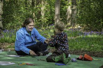 A woman sits on a green tarpaulin on the woodland floor as she interacts with a young child