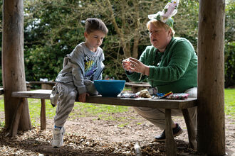 A woman in a green jumper wearing bunny ears helps a young boy make a bird feeder in an outdoor setting