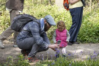 A mean kneels down to show his young daughter how to take a photo of bluebells on his phone